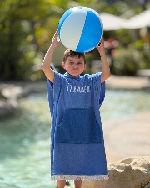 Child in a blue Hello Stranger poncho towel holding a beach ball beside the water